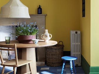 Dining space with brigh yellow walls and green skirting boards alongside a wooden table and blue stool.