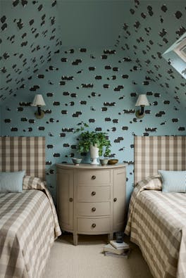 Children's bedroom featuring pale blue dog and sheep print wallpaper (Nip & Lassie - Celestial Blue) with two beds and a bedside drawer in the middle.