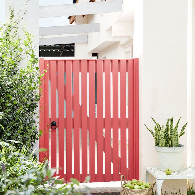 Coral red garden gate painted in 'Orange Aurora', alongside neutral white walls in 'Hollyhock', plants and bushes.