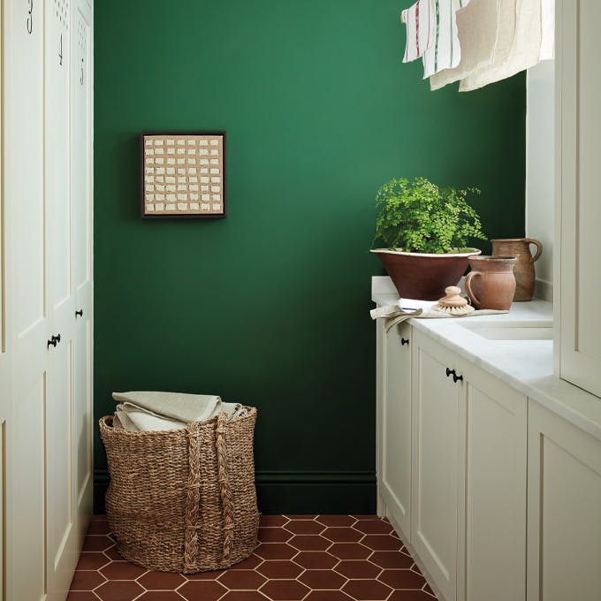 Laundry room painted in dark green shade 'Dark Brunswick' with a laundry basket, white cupboards and tiled floor. 