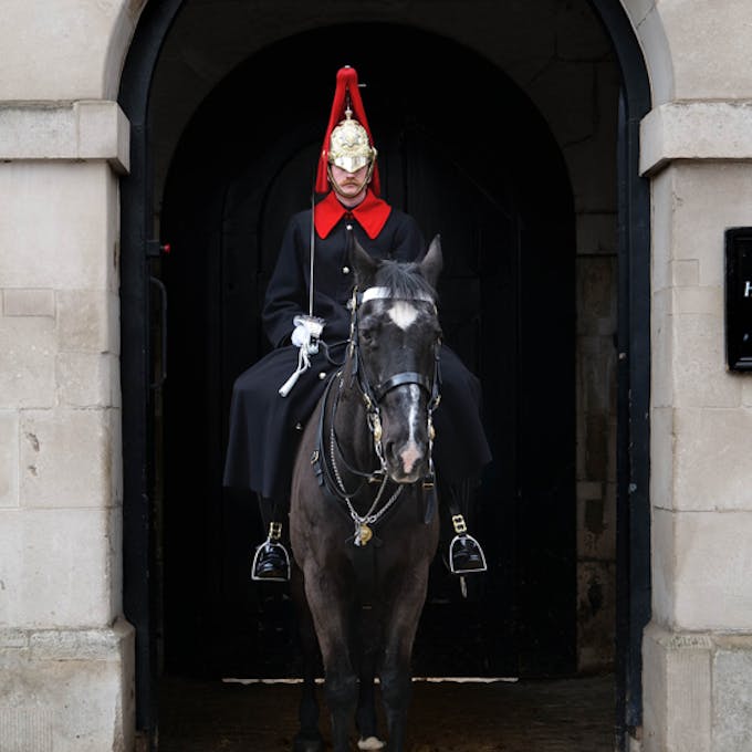 A guard wearing a long black coat with a red collar and a gold and red helmet sitting on a horse under an archway.