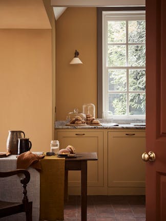 Kitchen space painted in honey shade 'Bombolone' with a paneled window, wooden table and chair.