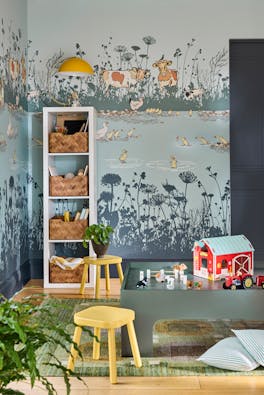 Children's playroom featuring blue mural wallpaper (Riverside Capers - Moo) with storage space to the left alongside yellow stools and toys in the foreground.