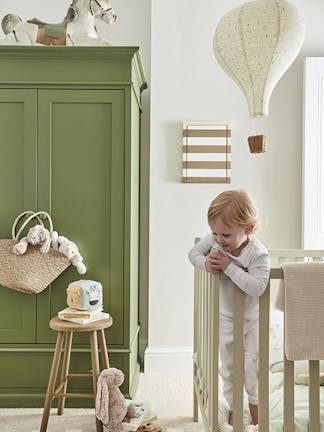 Calm nursery space with a green (Green Stone - Pale) wardrobe on the left and a crib on the right with a side table and fluffy toys on the floor.