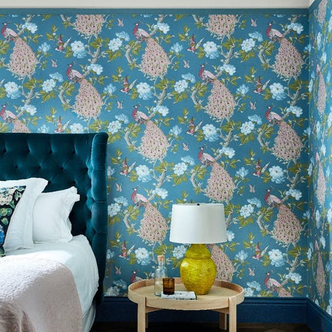 Bedroom featuring blue wallpaper with a vibrant peacock print (Pavona – Sylvie), behind a bed with a green velvet headboard.