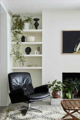 Living space painted in warm neutral Slaked Lime - Mid with shelves containing plants and vases behind a black leather chair.