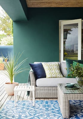 Outdoor living space painted in the dark green shade, 'Mid Azure Green' behind a weave sofa, table and plant.