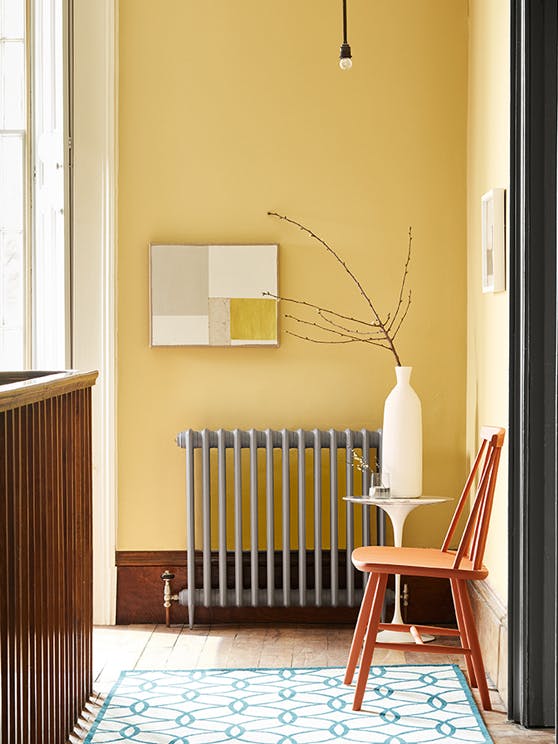 Landing decorated with Light Gold walls, grey radiator, an orange chair, a patterned rug, and a white side table and vase.