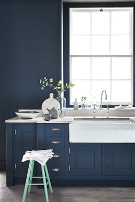 Kitchen painted in dark blue (Hicks' Blue) with a contrasting bright green (Green Verditer) stool underneath a window.