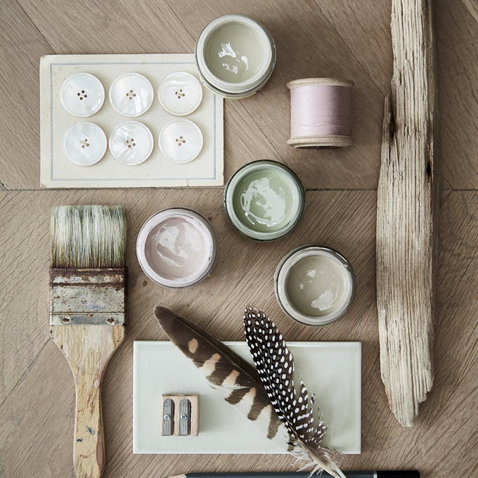 Close-up flat lay of a paintbrush, buttons, feathers, thread, and three paint sample pots in light pink, green, and grey.