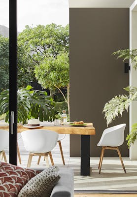 Outside dining space with a wooden table and white chairs surrounded by greenery and a high wall to the right painted in grey-brown shade 'Attic II'.