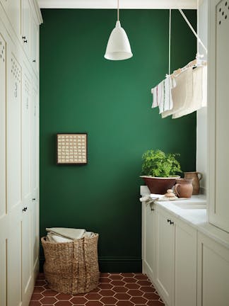 Laundry room painted in dark green shade 'Dark Brunswick' with a laundry basket, white cupboards and tiled floor. 