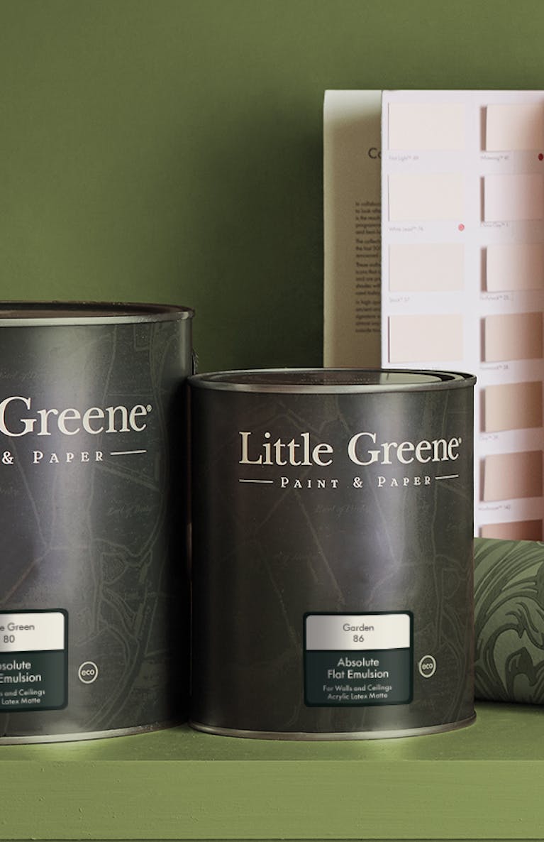 A shelf and wall painted in green (Garden) with two paint cans, an open colour card, a roll of floral green wallpaper (Poppy Trail - Sage Green) and a small plant in a round vase on top of the shelf.