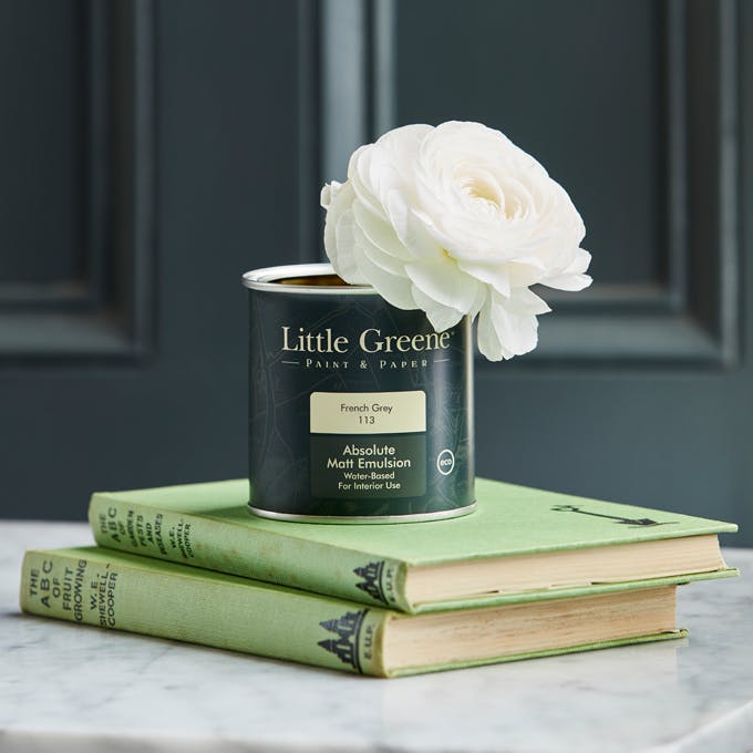 Close-up of a black Little Greene paint tin with a white flower inside, placed on two green books, in front of a black wall.