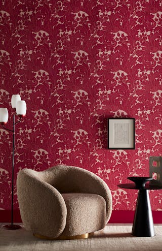 Close-up of a living space featuring red scroll wallpaper (Tulip - Theatre), a fluffy gray armchair, a side table and a lamp.