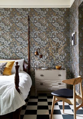 Bedroom with a paisley wallpaper (Pomegranate - Prophet) and a four poster bed with drawers and a black chair.