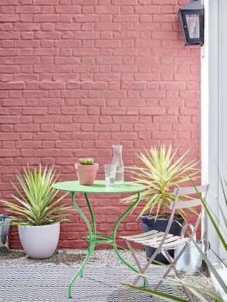 Outside brick wall painted in red shade 'Ashes of Roses' with bright green garden table and pink chair.