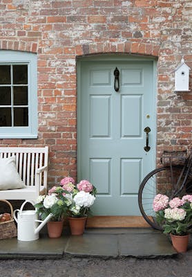 Front door painted in grey blue shade 'Celestial Blue'  with a brick surround and a grey bench with flowers.