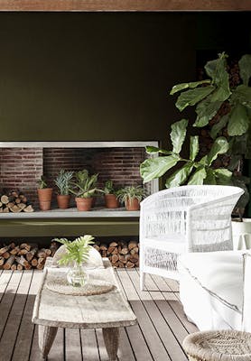 Outside living area with dark green (Invisible Green) back wall and white chairs next to wooden table, surrounded by plants.