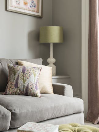 Living room corner featuring warm grey walls, grey sofa with cushions, a side table and a table lamp with a green shade.