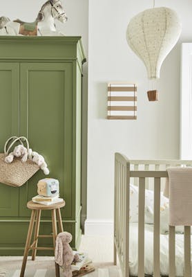 Calm nursery space with a green (Green Stone - Pale) wardrobe on the left and a crib on the right with a side table and fluffy toys on the floor.