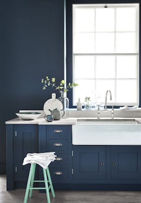 Kitchen painted in dark blue (Hicks' Blue) with a contrasting bright green (Green Verditer) stool underneath a window.