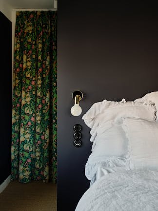 Bedroom wall painted with dark blue (Dock Blue) with a wall-mounted light, bed with white bedding and a colourful curtain.