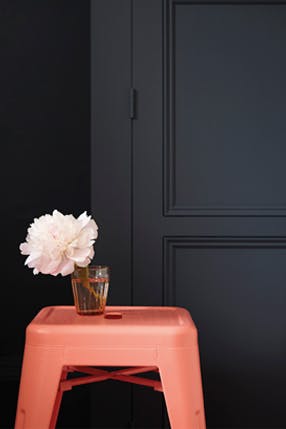 Bedroom painted in blue black 'Basalt' with a contrasting bright orange stool and a vase with a flower on top.