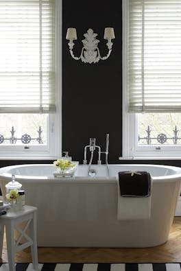 Bathroom with a freestanding bathtub and a black wall featuring two large windows and a chandelier wall light in between.