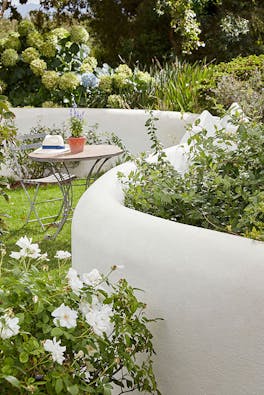 Garden area with a winding stone wall in light grey (Cool Arbour) and table and chairs in the background alongside greenery.