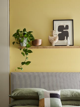 Bed with cushions below a wall painted in vibrant yellow Sunlight, with a shelf topped with a plant, frame and ornaments.