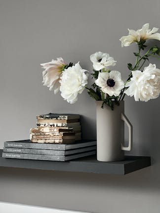 Close-up of a bookshelf holding books stacked on top of each other and white flowers in a grey vase against a matching wall.