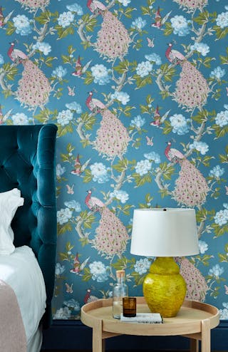 Bedroom featuring blue wallpaper with a vibrant peacock print (Pavona – Sylvie), behind a bed with a green velvet headboard.