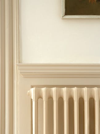 Close-up of a cast iron radiator painted in neutral beige (Stock - Dark), in front of a coordinating architrave and panel.