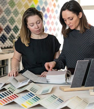 Two women looking through Little Greene's Color Cards on a table next to a wall comprised of multi-colored cubes.