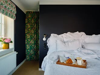 A bedroom with dark Dock Blue walls, a large bed covered with plush white bedding and a colorful floral curtain.