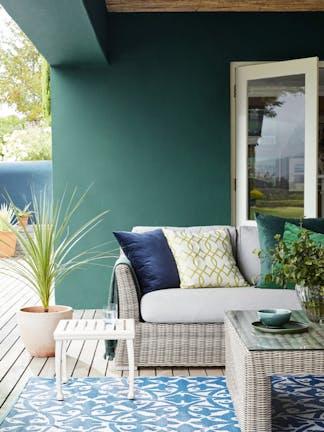 Outdoor dining area with dark green wall (Mid Azure Green) and wicker sofa with table and contrasting blue patterned rug.