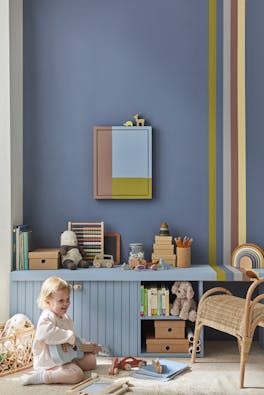 Child's playroom painted in dark blue (Juniper Ash) with multi colored stripes on the right, and toys placed on top of a unit with a child playing guitar.