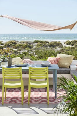 Beachside balcony with a dining table painted in muted blue 'Etruria' and chairs in lime green 'Pale Lime' overlooking the ocean and greenery.