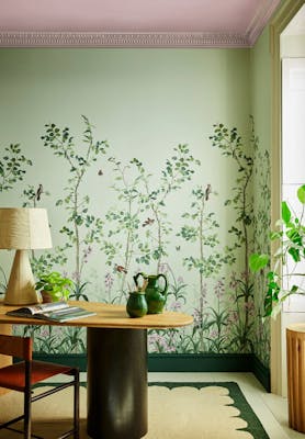 Green floral mural wallpaper featuring birds and butterflies (Bird & Bluebell - Pea Green) with a lamp and plant on a desk.