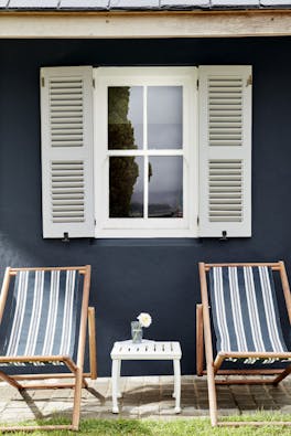 Two deck chairs in front of an exterior window with light grey shutters and a wall painted in deep black blue 'Basalt'.