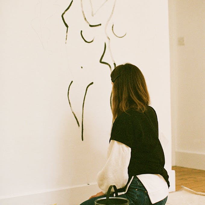 A female artist painting the outline of a human body with dark green paint on an interior white wall as a part of a mural.