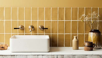 A white sink on a wooden counter with wall mounted taps and spout in front of a backsplash utilising Bassoon yellow tiles.