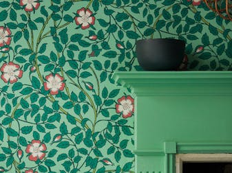 Close-up of floral wallpaper (Briar Rose - Green Verditer) with the corner of a green fireplace surround in front.