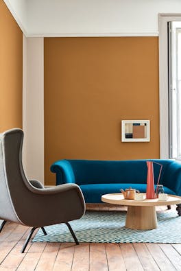 Living space painted in dark orange (Middle Buff) with a contrasting deep blue sofa, grey armchair and wooden table on a rug.