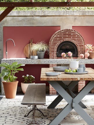 Outdoor kitchen painted in deep terracotta red 'Tuscan Red' with brickwork, plants and a wooden table and bench.