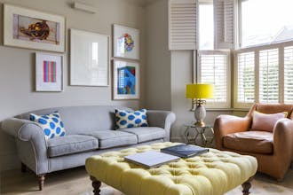 Living room scheme with grey walls, a bay window with white blinds, a sofa, an armchair and a large buttoned footstool.