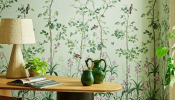 National Trust green floral mural wallpaper featuring birds and butterflies (Bird & Bluebell - Pea Green) behind a wood desk.