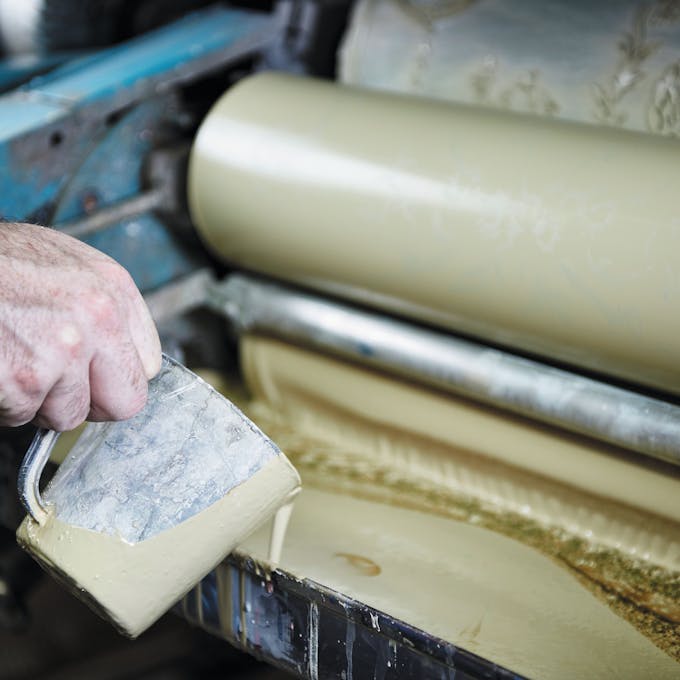 Close-up of a hand pouring beige paint into a factory machine.