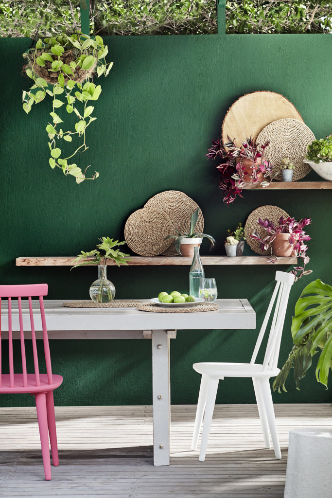 Outdoor dining space featuring a dark green (Puck) wall, a light grey (French Grey) table and a bright pink (Carmine) chair.